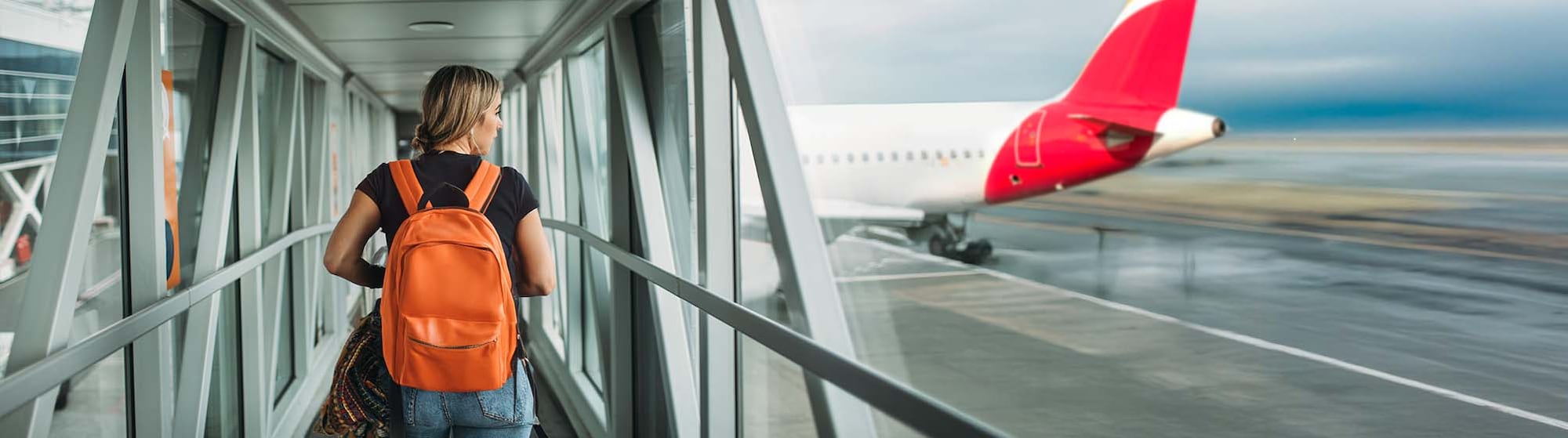 The image shows a woman with an orange backpack walking across a pedestrian bridge at an airport. There is also an airplane in the background.