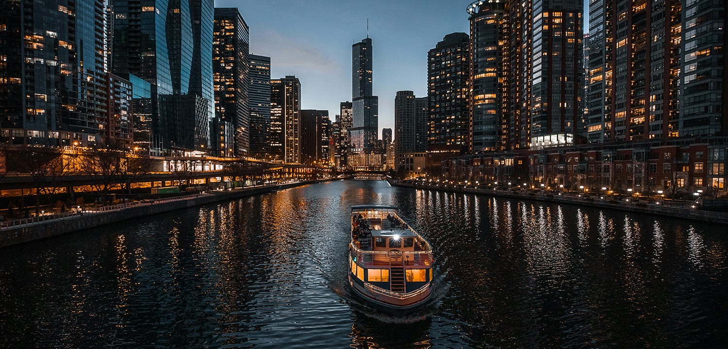 A boat sails along the Chicago River, surrounded by impressive skyscrapers at sunset