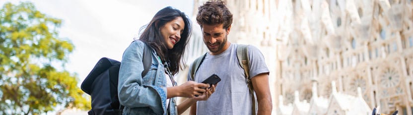 La imagen muestra a una pareja mirando un teléfono móvil, en el fondo aparece un monumento
