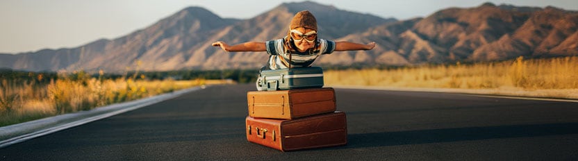 La imagen muestra a un niño viajando en una caja de cartón por una carretera desértica con montañas al fondo, simulando un avión en vuelo.