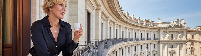 Una mujer disfrutando de un café en una terraza con vistas a un edificio histórico y arcos monumentales.
