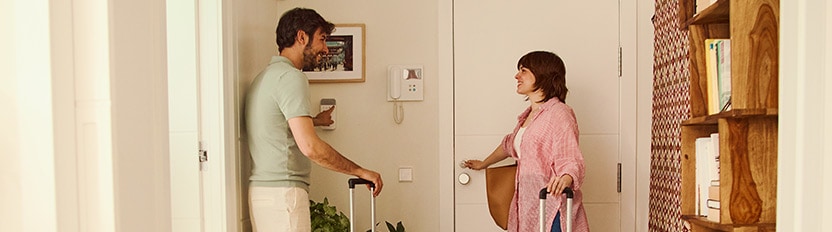 La imagen muestra a una pareja saliendo por la puerta de su casa mientras portan dos maletas