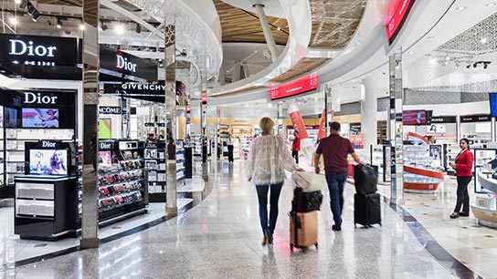 La imagen muestra a una pareja caminando con su equipaje por un area Duty Free del aeropuerto 